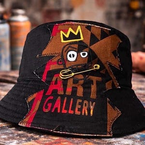 May include: A black bucket hat featuring a brown patch with a yellow crown graphic, a stylized face, and the text ART GALLERY in red and yellow letters. A gold-toned safety pin is attached to the front of the hat over the face graphic.