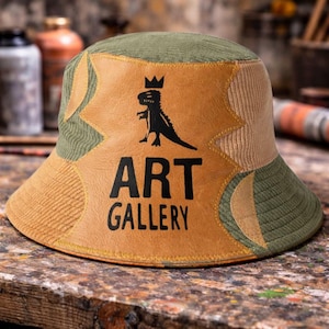 May include: A patchwork bucket hat with a dinosaur graphic and the words "ART GALLERY" in black. The hat features olive green, tan, and brown corduroy panels. The dinosaur is wearing a crown.