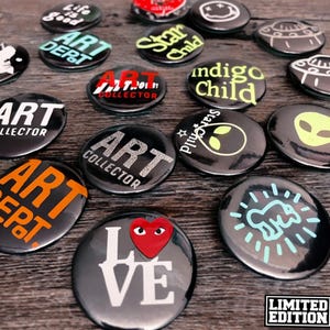 May include: A collection of 15 round black and white buttons with various designs. The designs include text, logos, and illustrations. Some of the text includes "Art Dept", "Art Collector", "Star Child", "Indigo Child", "Love", and "Limited Edition".