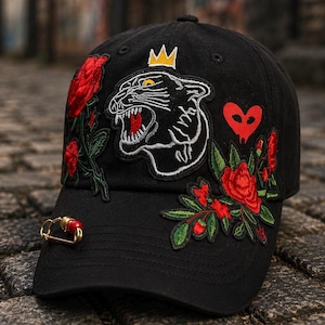 May include: Black baseball cap with embroidered patches of a panther, roses, and a heart. The cap features a gold-colored safety pin with a red bead. The crown of the cap has a small gold crown.