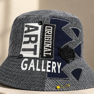 Może przedstawiać: Szary sztruksowy kapelusz typu bucket hat z białą naszywką z napisem ART GALLERY, czarną naszywką z napisem ORIGINAL, ciemnoniebieskim panelem z trójkątnymi przeszyciami, czarną aplikacją róży oraz żółtą agrafką jako akcentem.