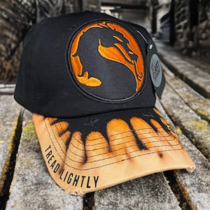 May include: Black baseball cap with an orange dragon logo on a circular patch. The brim is tan with a burnt orange design and the words "TREAD LIGHTLY" printed on it. A black tag hangs from the side.