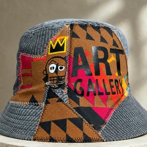 May include: A grey corduroy bucket hat featuring a large, multi-colored fabric patch on the front. The patch displays a stylized face with a yellow crown, the text ART GALLERY in black, and geometric patterns in brown, black, and pink.
