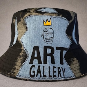 Puede incluir: Un sombrero de pescador negro con un patrón desgastado y desteñido, que presenta un parche central de mezclilla azul claro. El parche muestra un rostro dibujado a mano con una corona amarilla encima y el texto negro ART GALLERY en negrita.