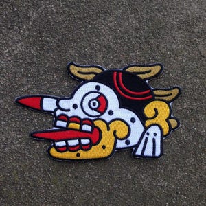 以下が含まれることがあります： スタイリッシュでカラフルなデザインの刺繍パッチ。黒と赤のアクセント、長い赤い鼻、黄色の角を持つ白い顔が特徴です。デザインは黒と黄色で縁取られています。