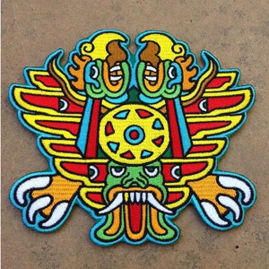 Puede incluir: Un parche bordado colorido que presenta una criatura mítica con alas, un diseño circular en su cabeza y una boca grande abierta con dientes afilados. La criatura está representada en una paleta vibrante de rojo, amarillo, verde, azul y blanco.