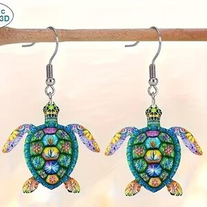 May include: A pair of colorful acrylic turtle earrings. Each earring features a detailed turtle design with blue, green, yellow, and purple accents. The earrings are suspended from silver-colored fish hook ear wires. The text "2D Acrylic Flat NOT 3D" is visible.