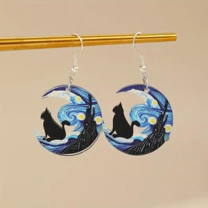 May include: A pair of earrings featuring a black cat silhouette perched on a crescent moon. The moon design incorporates a swirling blue and white starry night sky, with a dark tree silhouette at the bottom. The earrings hang from silver hooks.