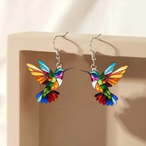 May include: A pair of colorful hummingbird earrings with silver hooks. The hummingbirds are made of enamel and have a stained glass look. The earrings are a vibrant mix of red, orange, yellow, green, blue, and purple.