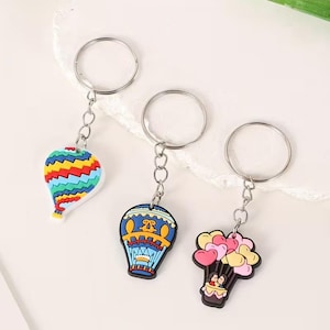 May include: Three colorful hot air balloon keychains. One is striped with red, yellow, blue, and green. The second is blue with a yellow and orange design. The third features heart-shaped balloons in pink and yellow.