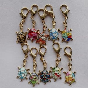 May include: A collection of gold-toned turtle charms with lobster claw clasps. The turtle shells feature various colorful designs, including leopard print, floral patterns, and geometric shapes. These charms are suitable for jewelry making or crafting.