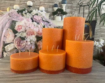 May include: Four orange pillar candles with a textured surface and a darker orange band around the bottom. The candles are arranged in a row on a wooden surface.
