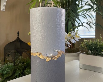 May include: A gray and white pillar candle with gold leaf accents. The candle has a textured surface and is decorated with small white beads.