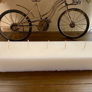 Puede incluir: Una vela rectangular blanca con cuatro mechas, sentada sobre una superficie de madera. La vela está lista para ser encendida.