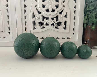 May include: Four green sphere candles of varying sizes, with a textured surface, arranged in a row on a white wooden surface.