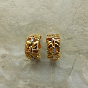 Peut inclure: Une paire de boucles d'oreilles créoles dorées avec un motif de papillon. Les boucles d'oreilles ont une surface texturée et un accent argenté au centre. Les boucles d'oreilles sont présentées sur une surface gris clair.