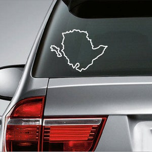 May include: White outline of a geographical region sticker applied to the rear window of a silver vehicle. The sticker depicts the shape of a landmass with a smaller adjacent island, likely a state or country outline.