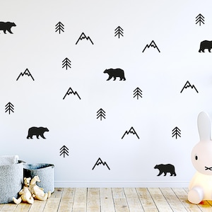 Könnte beinhalten: Eine weiße Wand mit schwarzen Bären-, Berg- und Baumaufklebern. Der Raum hat einen Holzboden, zwei graue Aufbewahrungskörbe und eine hasenförmige Lampe. Zwei Stofftiere befinden sich vor den Körben.