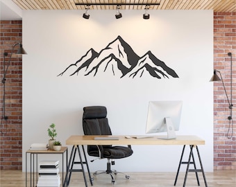 May include: A black mountain range wall decal is mounted on a white wall above a desk. The office space includes a black office chair, a wooden desk, and a small side table with books and a plant. The wall is partially brick.