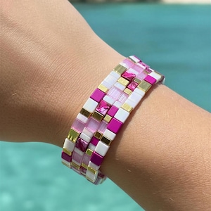 Puede incluir: Una pulsera colorida con un diseño de mosaico. La pulsera presenta pequeñas baldosas cuadradas en tonos de rosa, blanco y dorado. La pulsera se usa en una muñeca, con un fondo de agua turquesa.