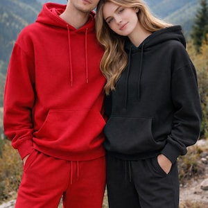 K&ouml;nnte beinhalten: Ein Paar in passenden Kapuzenpullovern und Jogginghosen. Eine Person tr&auml;gt einen roten Kapuzenpullover und eine Jogginghose, die andere Person tr&auml;gt Schwarz. Beide Outfits haben einen Kordelzug und eine Vordertasche. Im Hintergrund ist ein Gebirge zu sehen.