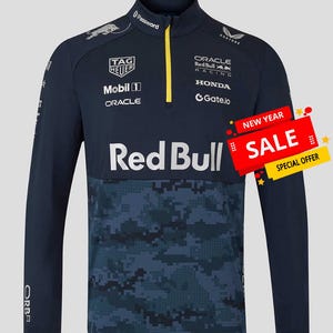 Puede incluir: Camiseta de manga larga azul marino con cuello con media cremallera y un patrón de camuflaje en la mitad inferior. La camiseta presenta múltiples logotipos, incluyendo "Red Bull", "TAG Heuer" y "Oracle". Se ve una pancarta de "SALE". La camiseta está diseñada para ropa deportiva.