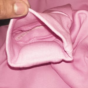 K&ouml;nnte beinhalten: Nahaufnahme eines rosafarbenen Sweatshirt-&Auml;rmelb&uuml;ndchens. Das B&uuml;ndchen ist gerippt und zur&uuml;ckgefaltet, wodurch das Innenfutter sichtbar wird. Der Stoff hat eine einfarbige, weiche rosa Farbe. Das Bild konzentriert sich auf die Textur und Verarbeitung des Kleidungsst&uuml;cks.
