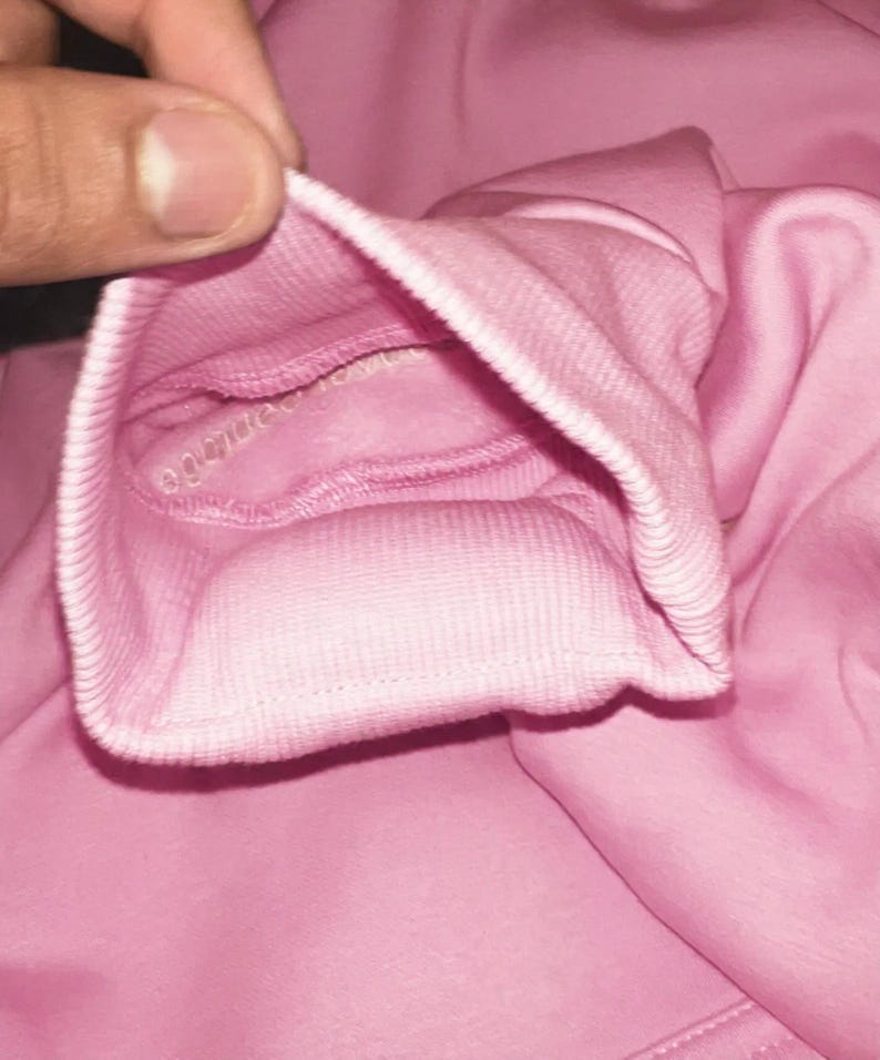 K&ouml;nnte beinhalten: Nahaufnahme eines rosafarbenen Sweatshirt-&Auml;rmelb&uuml;ndchens. Das B&uuml;ndchen ist gerippt und zur&uuml;ckgefaltet, wodurch das Innenfutter sichtbar wird. Der Stoff hat eine einfarbige, weiche rosa Farbe. Das Bild konzentriert sich auf die Textur und Verarbeitung des Kleidungsst&uuml;cks.