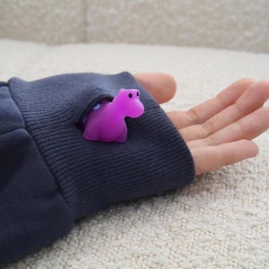 K&ouml;nnte beinhalten: Eine kleine, lila Nilpferdfigur schaut aus einem marineblauen &Auml;rmel hervor. Das Nilpferd hat schwarze Augen. Der &Auml;rmel ist dunkelblau und hat ein geripptes B&uuml;ndchen. Der Hintergrund ist eine helle, strukturierte Oberfl&auml;che.