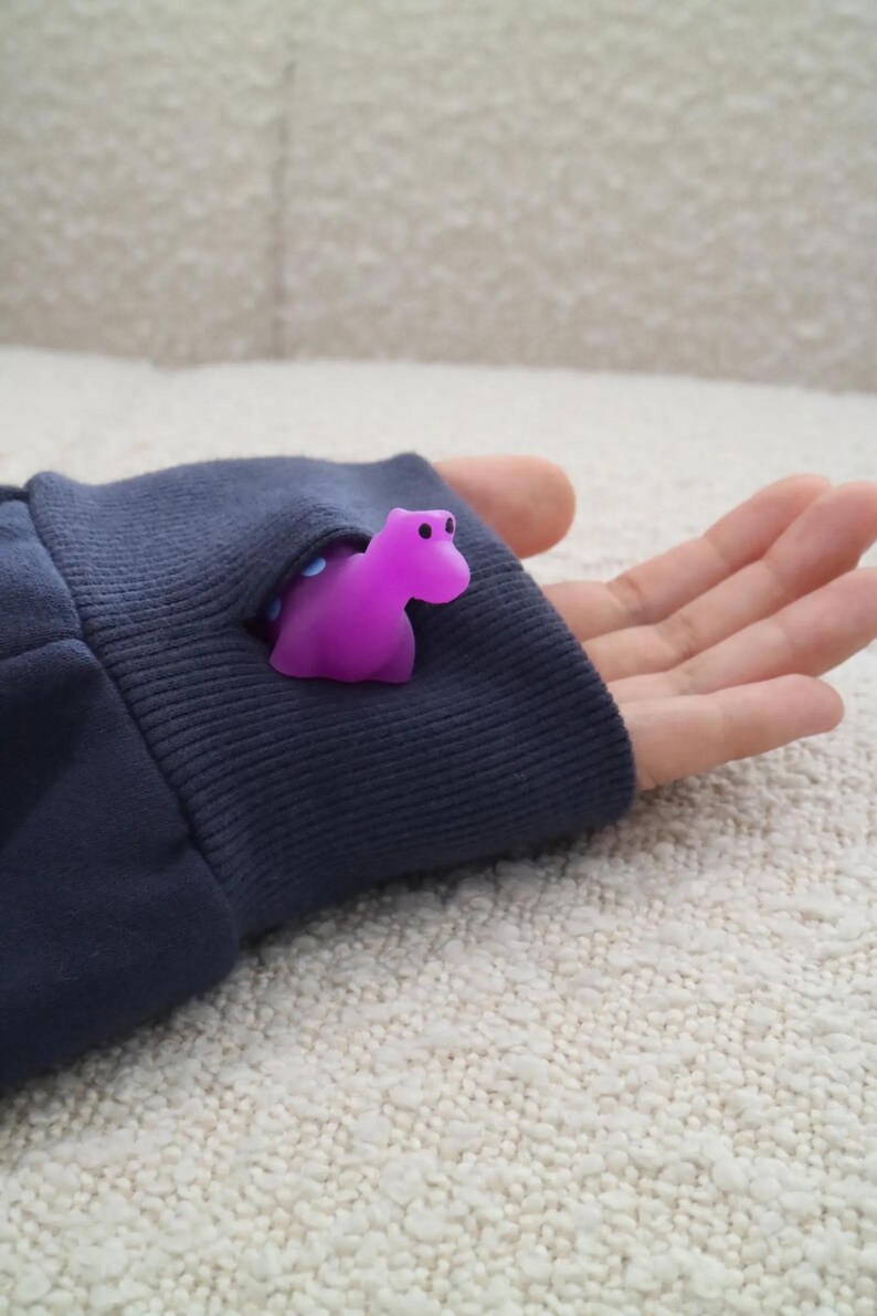 K&ouml;nnte beinhalten: Eine kleine, lila Nilpferdfigur schaut aus einem marineblauen &Auml;rmel hervor. Das Nilpferd hat schwarze Augen. Der &Auml;rmel ist dunkelblau und hat ein geripptes B&uuml;ndchen. Der Hintergrund ist eine helle, strukturierte Oberfl&auml;che.