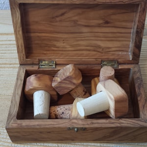 Può includere: Una scatola di legno con coperchio a cerniera, riempita con diversi tappi per bottiglie in legno. I tappi hanno una finitura in legno naturale e una base in sughero. La scatola e i tappi sono in legno d'ulivo.