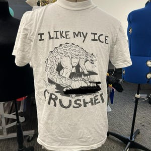 Könnte beinhalten: Ein weißes T-Shirt mit dem Text "I LIKE MY ICE" über einer Schwarz-Weiß-Illustration eines Dinosauriers, der Eis zermalmt. Das Wort "CRUSHER" ist unter der Illustration in einer verwaschenen Schrift gedruckt.