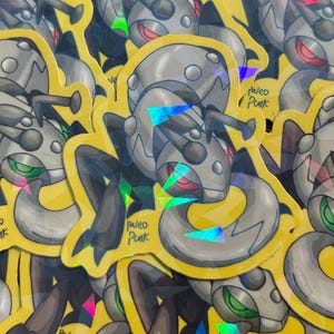 May include: A holographic sticker featuring a stylized metallic gray insectoid creature with dark limbs and glowing red eyes. The design includes a yellow border and the text Paleo Punk written in a black script font on the lower left side.
