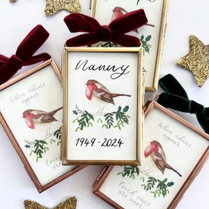 May include: A collection of framed memorial ornaments with a robin illustration, text, and velvet bows. The frames are gold and copper-toned, with text that reads "Grandpa" and "Nanny". Gold glitter stars and green velvet bows are also present.