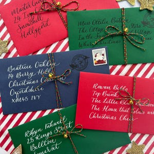 Pode incluir: Cinco envelopes com tema natalino com endere&ccedil;os escritos &agrave; m&atilde;o e decora&ccedil;&otilde;es festivas. Cada envelope &eacute; amarrado com barbante e um pequeno sino. Os envelopes est&atilde;o dispostos em um fundo listrado vermelho e branco.