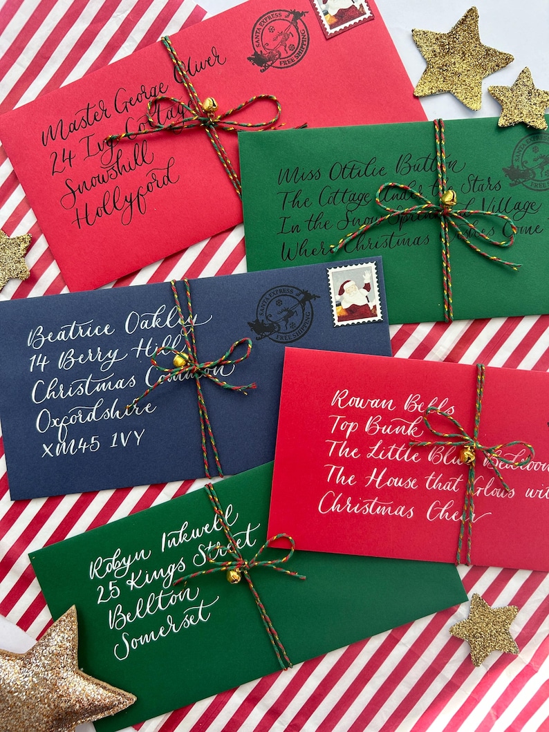 Pode incluir: Cinco envelopes com tema natalino com endere&ccedil;os escritos &agrave; m&atilde;o e decora&ccedil;&otilde;es festivas. Cada envelope &eacute; amarrado com barbante e um pequeno sino. Os envelopes est&atilde;o dispostos em um fundo listrado vermelho e branco.