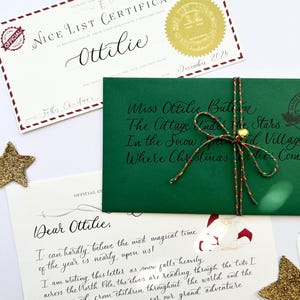 Pode incluir: Um envelope verde com um selo dourado e uma corda vermelha e branca amarrada ao redor. O envelope &eacute; endere&ccedil;ado a Miss Ottilie Button, The Cottage Under the Stars, In the Snow,  Spiced Village, Where Christmas Wishes Come. Uma carta escrita &agrave; m&atilde;o com um desenho do Papai Noel e um duende &eacute; vis&iacute;vel na mesa ao lado do envelope.