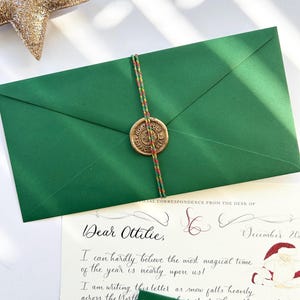 Pode incluir: Um envelope verde com um selo de lacre e uma carta manuscrita endere&ccedil;ada a Ottilie. A carta diz: "Querida Ottilie, Mal posso acreditar que a &eacute;poca mais m&aacute;gica do ano est&aacute; quase chegando! Estou escrevendo esta carta enquanto a neve cai forte no Norte..."