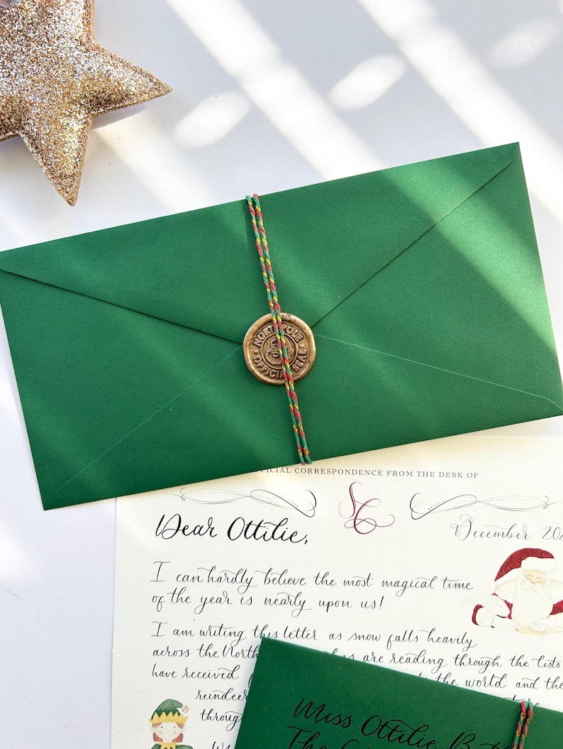 Pode incluir: Um envelope verde com um selo de lacre e uma carta manuscrita endere&ccedil;ada a Ottilie. A carta diz: "Querida Ottilie, Mal posso acreditar que a &eacute;poca mais m&aacute;gica do ano est&aacute; quase chegando! Estou escrevendo esta carta enquanto a neve cai forte no Norte..."