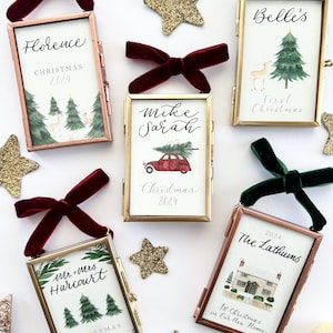 Può includere: Ornamenti natalizi in cornici rettangolari con illustrazioni ad acquerello e testo, ciascuno con un fiocco di velluto. Stelle e campane dorate glitterate sono sparse. Il testo include nomi e l'anno 2024. Cornici in oro e rame.