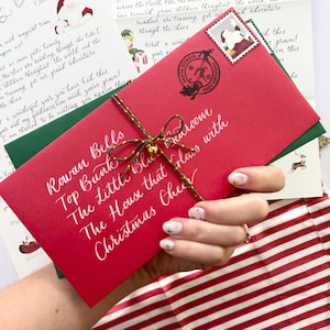 Pode incluir: Um envelope vermelho com caligrafia branca que diz "Rowan Bells Top Bunk The Little Blue Bedroom The House that Claus with Christmas Cheer". O envelope est&aacute; amarrado com um cord&atilde;o dourado e um amuleto de sino dourado.