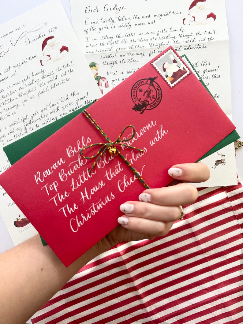 Pode incluir: Um envelope vermelho com caligrafia branca que diz "Rowan Bells Top Bunk The Little Blue Bedroom The House that Claus with Christmas Cheer". O envelope est&aacute; amarrado com um cord&atilde;o dourado e um amuleto de sino dourado.