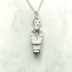 May include: A white porcelain pendant necklace featuring a small figurine of a person with black hair, a black bow, and black shoes. The pendant is attached to a silver chain.