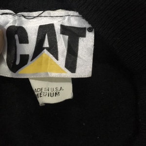 May include: A white tag with black text that reads "CAT" in a bold, sans-serif font. A yellow triangle is behind the text. The tag also has a smaller white tag attached to it that reads "MADE IN U.S.A. MEDIUM".