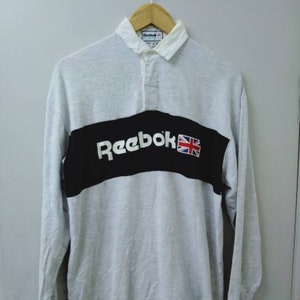 Op de afbeelding: Een wit rugby shirt met lange mouwen en een zwarte band over de borst. De band heeft het woord "Reebok" in witte letters en een kleine Union Jack-vlag.