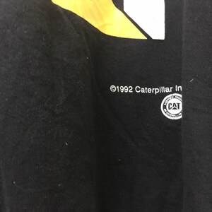 May include: Black t-shirt with a yellow and white graphic design. The text "&copy;1992 Caterpillar Inc." is printed on the bottom of the shirt. A circular logo with the text "Caterpillar Inc." and "CAT" is printed on the bottom right of the shirt.