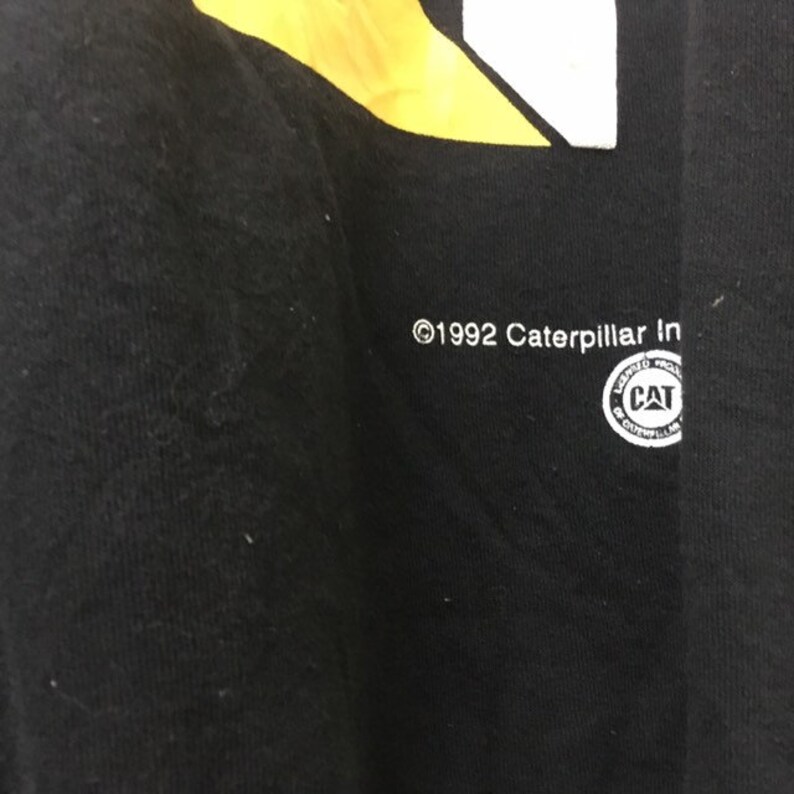 May include: Black t-shirt with a yellow and white graphic design. The text "&copy;1992 Caterpillar Inc." is printed on the bottom of the shirt. A circular logo with the text "Caterpillar Inc." and "CAT" is printed on the bottom right of the shirt.