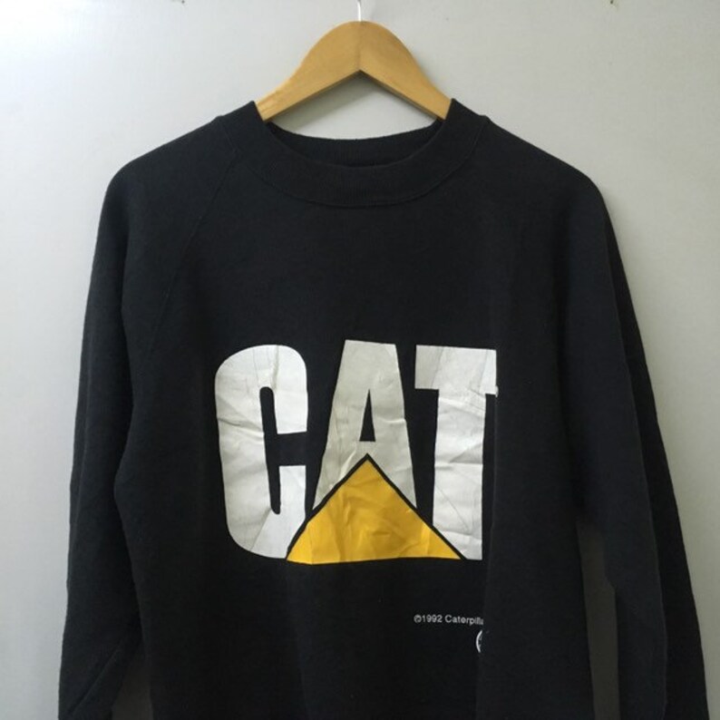 May include: Black sweatshirt with a white "CAT" logo and a yellow triangle. The logo is printed on the front of the sweatshirt. The text "&copy;1992 Caterpillar" is printed below the logo.