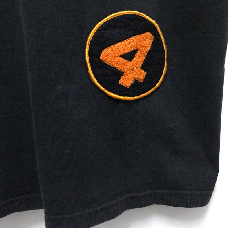 Peut inclure: T-shirt noir avec un grand patch brod&eacute; circulaire orange et noir repr&eacute;sentant le chiffre "4" dans une police stylis&eacute;e.
