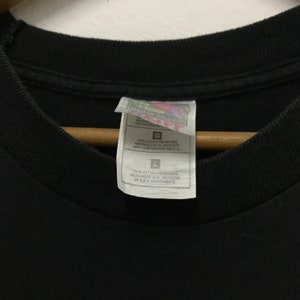 Peut inclure: Une &eacute;tiquette de t-shirt noire avec du texte blanc qui indique "Field of the Loom" et "100% coton pr&eacute;-r&eacute;tr&eacute;ci assembl&eacute; au Salvador avec des composants des &Eacute;tats-Unis."